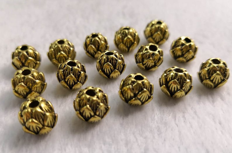 May include: A collection of small, round, gold-toned metal beads, each featuring a textured, layered petal design resembling an artichoke or lotus flower. Each bead has a central hole for stringing, with a dark, antiqued finish in the crevices.