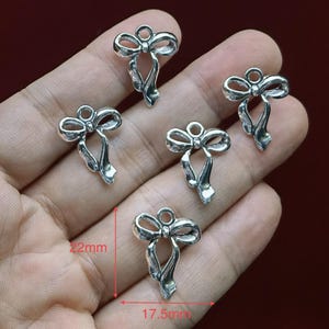 Puede incluir: Cinco dijes de metal plateado con forma de lazo y una anilla en la parte superior para su fijación. Los dijes presentan un diseño de cinta detallado. Un indicador de medida rojo muestra una altura de 22 mm y una anchura de 17,5 mm.