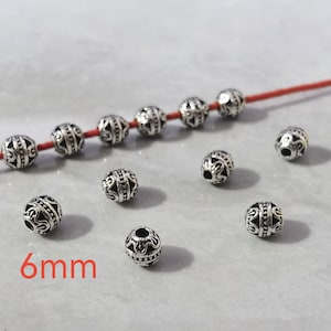 May include: A collection of small, round, silver-toned metal beads with intricate embossed patterns and a central horizontal band. Six beads are threaded onto a thin red cord, while five additional beads are scattered nearby. The text 6mm is visible.