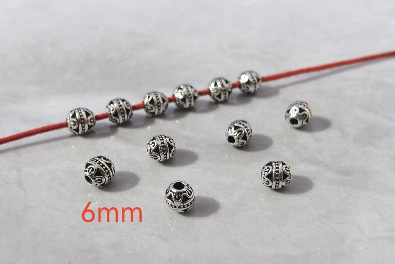 May include: A collection of small, round, silver-toned metal beads with intricate embossed patterns and a central horizontal band. Six beads are threaded onto a thin red cord, while five additional beads are scattered nearby. The text 6mm is visible.