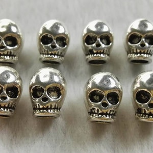 May include: Eight small, metallic silver-colored skull beads arranged in two rows of four. Each bead features a polished, reflective surface with recessed, darkened details defining the eye sockets, nasal cavity, and teeth along the lower jaw.