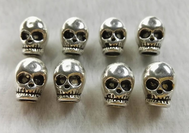 May include: Eight small, metallic silver-colored skull beads arranged in two rows of four. Each bead features a polished, reflective surface with recessed, darkened details defining the eye sockets, nasal cavity, and teeth along the lower jaw.