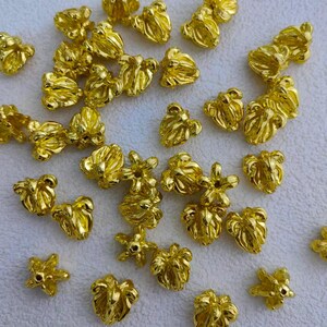 May include: A collection of small, gold-colored metal beads. The beads have a flower-like shape with multiple petals and a hole in the center. They are scattered on a white surface, creating a textured and shiny appearance. Ideal for jewelry making.