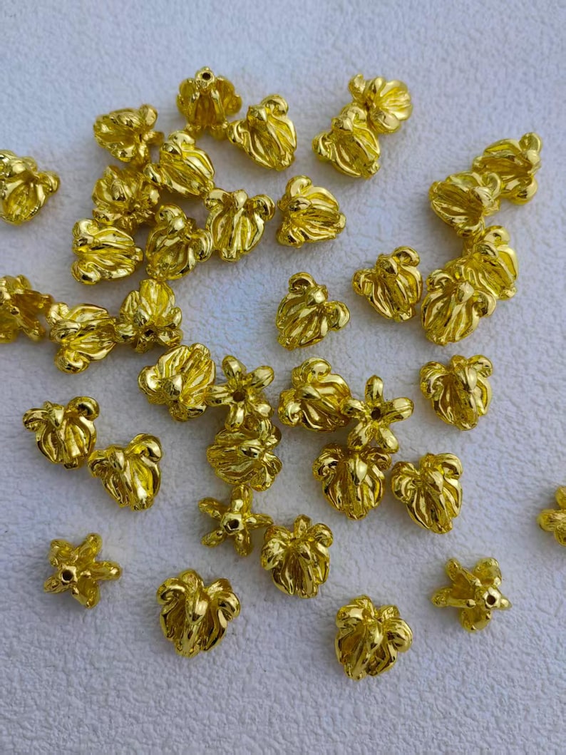 May include: A collection of small, gold-colored metal beads. The beads have a flower-like shape with multiple petals and a hole in the center. They are scattered on a white surface, creating a textured and shiny appearance. Ideal for jewelry making.