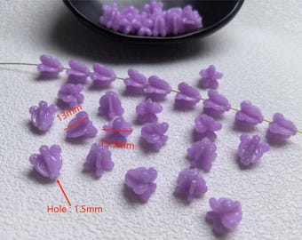 May include: Close-up of lavender-colored flower-shaped beads. The beads are approximately 13mm in length and 13.5mm wide, with a 1.5mm hole. Some beads are strung on a wire, while others are loose. A black dish holds more beads.