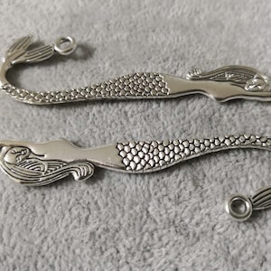 May include: Two silver mermaid bookmark charms with a detailed design. Each charm features a mermaid with a flowing tail and a textured, scaly pattern.