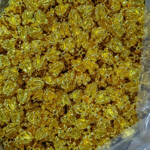 May include: A close-up shot of a large quantity of small, gold-colored, flower-shaped beads. The beads are clustered together, filling the frame. Each bead has a detailed, three-dimensional floral design, with multiple petals and a central opening. The beads have a shiny, metallic finish.