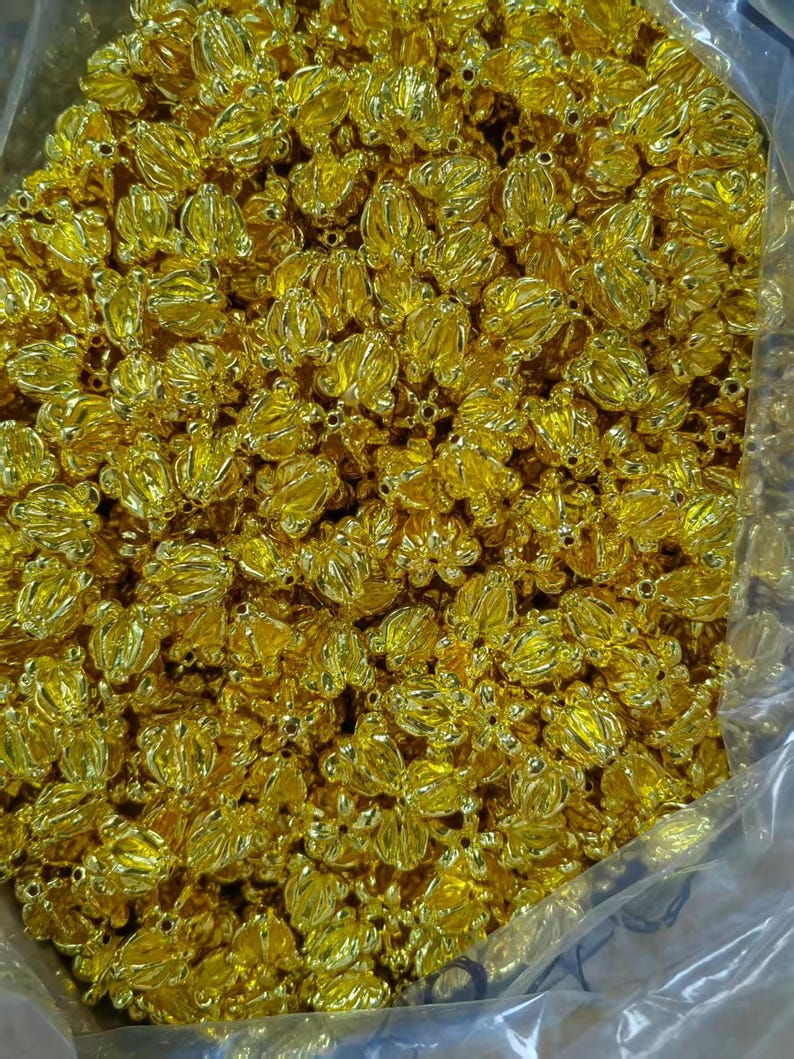 May include: A close-up shot of a large quantity of small, gold-colored, flower-shaped beads. The beads are clustered together, filling the frame. Each bead has a detailed, three-dimensional floral design, with multiple petals and a central opening. The beads have a shiny, metallic finish.