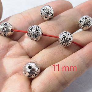 May include: Six spherical silver-toned metal beads with intricate cut-out filigree patterns and a central beaded band. Three beads are threaded on a thin red cord, while three others are loose. Each bead measures 11 mm in diameter.