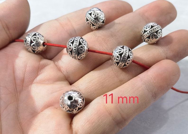 May include: Six spherical silver-toned metal beads with intricate cut-out filigree patterns and a central beaded band. Three beads are threaded on a thin red cord, while three others are loose. Each bead measures 11 mm in diameter.
