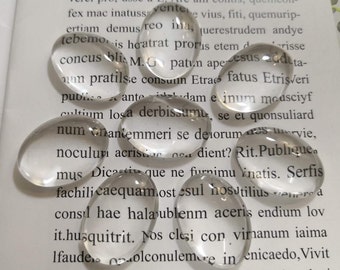 Pode incluir: Um close-up de vários cabochões ovais de vidro transparente, também conhecidos como gemas de vidro, em uma página de um livro. O livro está aberto e o texto é visível. As gemas de vidro são lisas e brilhantes.