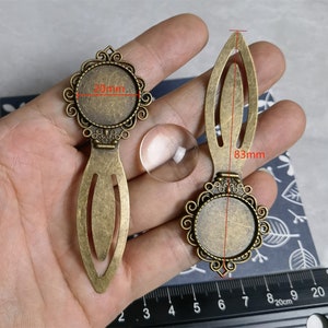 May include: Two antique bronze-colored metal bookmark blanks with ornate circular settings for cabochons, accompanied by a clear glass dome. The settings measure 20mm in diameter, and the total length of the bookmark is 83mm as shown by a ruler.
