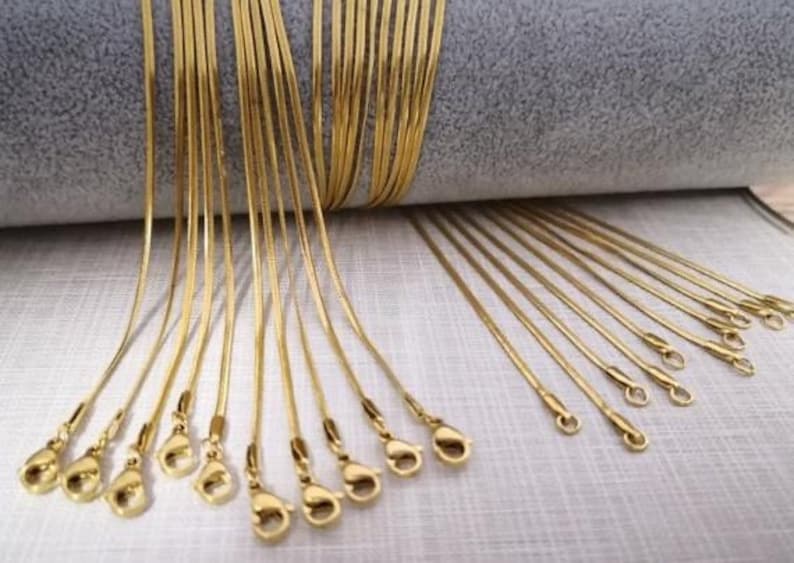 May include: A collection of gold snake chain necklaces with lobster claw clasps. The chains are laid out on a light gray surface.