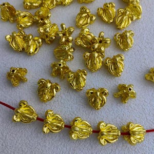 May include: A collection of small, shiny, gold-colored metal beads in a variety of floral shapes. Some beads are strung on a red cord. The beads have a detailed, textured surface and are scattered across a light background.
