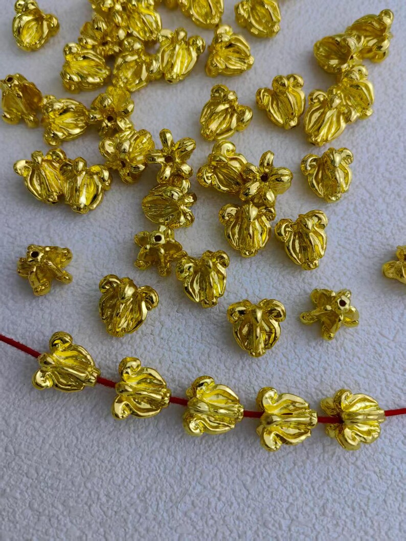 May include: A collection of small, shiny, gold-colored metal beads in a variety of floral shapes. Some beads are strung on a red cord. The beads have a detailed, textured surface and are scattered across a light background.