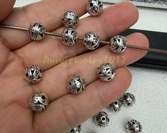 Puede incluir: Una colección de cuentas redondas de metal plateado con intrincados patrones de volutas en relieve. Cada cuenta mide 10 mm de diámetro con un orificio central de 2 mm. Varias cuentas están ensartadas en un alambre fino, mientras otras están sueltas.