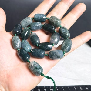 May include: A necklace of polished, barrel-shaped moss agate beads in shades of green and white. The beads are strung on a green cord, creating a natural aesthetic. The necklace is displayed on a light-coloured surface.