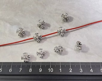 May include: Eight silver-toned metal beads shaped like ornate crosses, with one bead threaded onto a thin red cord. A black ruler is positioned at the bottom, showing measurements in centimetres, with the beads measuring approximately 1 cm in width.