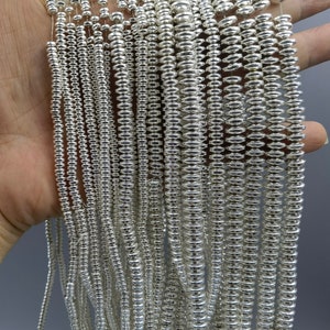 Könnte beinhalten: Ein Bündel silberfarbener Metallperlen, jede Perle ist ein kleiner Zylinder mit abgerundeter Ober- und Unterseite. Die Perlen sind in einem lockeren Haufen angeordnet, wobei sich einige Perlen überlappen.