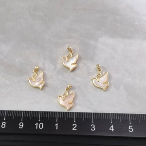 以下が含まれることがあります： 白い真珠貝の羽根を持つ、金色の鳩のチャームが4つ。チャームは、スケール用の定規のある白い表面に配置されています。