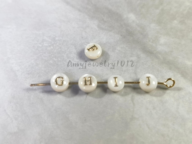 May include: Five round white beads with gold-colored letters, including one loose bead marked F and four beads on a gold-toned metal pin marked G, H, I, and J. The text Amyjewelry1012 is visible in the center of the image between the beads.