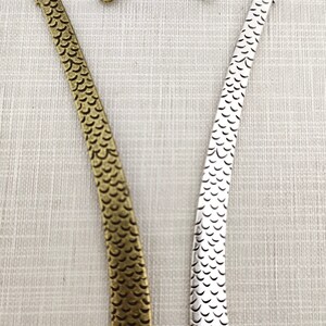 May include: Two metal bookmarks, one in an antique bronze finish and one in a bright silver finish. Each bookmark features a curved top shaped like a dragon head with a small circular loop, and a long, flat body textured with a repeating scale pattern.