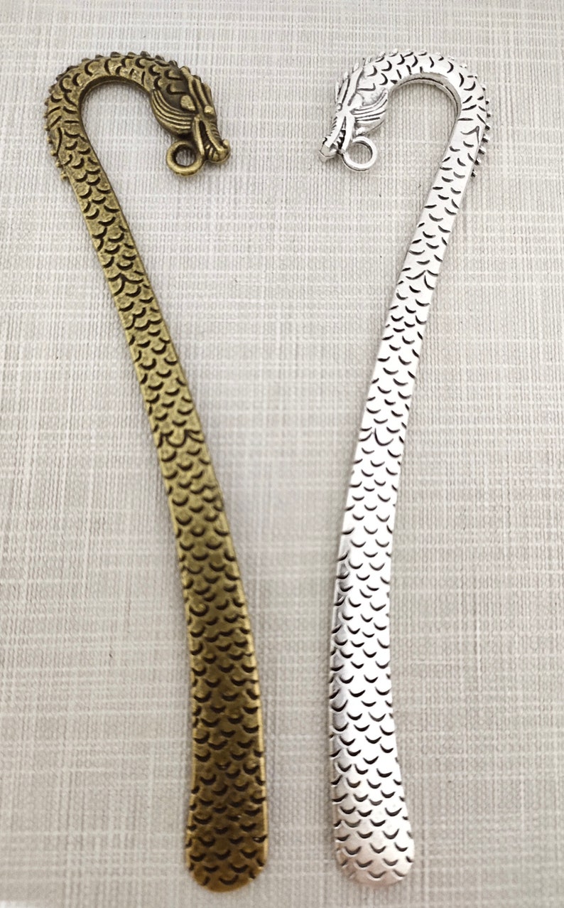 May include: Two metal bookmarks, one in an antique bronze finish and one in a bright silver finish. Each bookmark features a curved top shaped like a dragon head with a small circular loop, and a long, flat body textured with a repeating scale pattern.