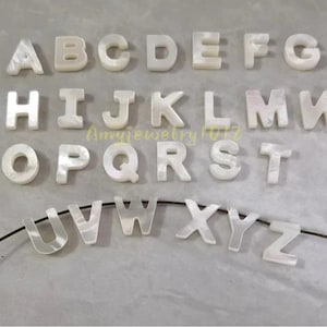 Puede incluir: Un conjunto completo de veintiséis letras del alfabeto de la A a la Z hechas de nácar blanco iridiscente. Cada letra presenta una superficie lisa y pulida con variaciones nacaradas naturales. Las letras de la U a la Z están ensartadas en un hilo negro fino.