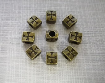 May include: Eight square-shaped metal beads with an antique bronze finish, each featuring a recessed cross design on the top surface. The beads are arranged in a circular pattern, with one bead placed in the centre showing a visible hole.