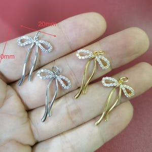 Peut inclure: Quatre pendentifs en forme de nœud ornés de pierres claires serties dans du métal. Deux pendentifs sont argentés et deux sont dorés. Chaque pendentif possède une petite boucle. Le texte rouge indique des dimensions de 20mm sur 30mm.