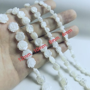 Könnte beinhalten: Nahaufnahme von Strängen weißer, rosenförmiger Perlen in den Größen 6 mm, 8 mm, 10 mm und 12 mm. Die Perlen sind aufgefädelt und bilden ein zartes Blumenmuster. Geeignet für die Schmuckherstellung oder Bastelprojekte.