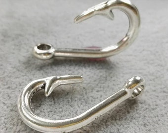 May include: Two silver-colored metal fish hook charms, each featuring a curved shank, a sharp barb, and a circular eyelet at the end for attachment. The charms have a smooth, polished finish and are positioned horizontally, showing their curved shape.