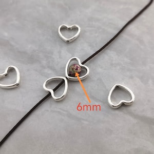 Peut inclure: Breloques en forme de cœur argentés avec une perle de 6 mm à motif floral au centre. Les breloques sont enfilées sur un cordon brun.