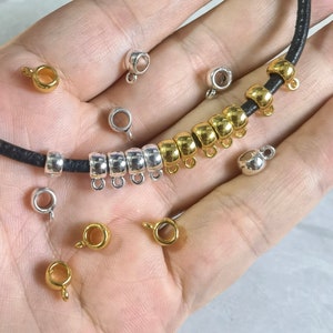 May include: A close-up of a hand holding a black leather cord with silver and gold metal bead charms. The cord is threaded with a variety of round beads, some silver and some gold. There are also several loose beads on the hand.