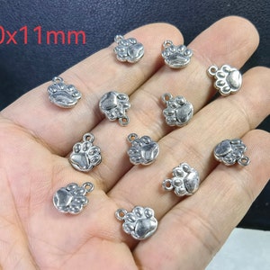 May include: A hand holding 10 silver paw print charms, each measuring 10x11mm. The charms have a loop at the top for attaching to jewellery or other crafts.