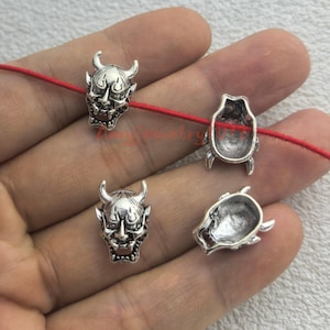May include: Four silver-toned metal beads shaped like Japanese Hannya masks. Two beads show the detailed front face with horns and fangs, while two show the hollow back. One bead is threaded onto a thin red cord, while the others are loose.
