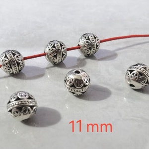 May include: Six spherical silver-toned metal beads with intricate cut-out patterns and a central band of small raised dots. Three beads are threaded on a thin red cord, while three others are loose. The text 11 mm is visible in red.