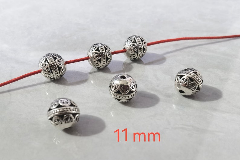 May include: Six spherical silver-toned metal beads with intricate cut-out patterns and a central band of small raised dots. Three beads are threaded on a thin red cord, while three others are loose. The text 11 mm is visible in red.