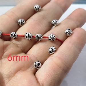 May include: A collection of round, silver-toned metal beads featuring intricate embossed patterns. Five beads are threaded onto a thin red cord, while several others are scattered loosely. The text 6mm is printed in red on the image.