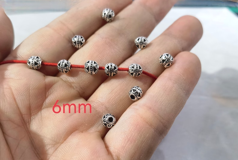 May include: A collection of round, silver-toned metal beads featuring intricate embossed patterns. Five beads are threaded onto a thin red cord, while several others are scattered loosely. The text 6mm is printed in red on the image.