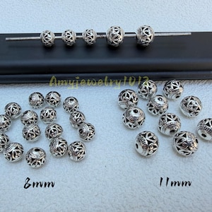 Puede incluir: Una colección de cuentas redondas de metal plateado con intrincados patrones de filigrana recortados. Las cuentas están dispuestas en dos grupos etiquetados como 8mm y 11mm, con cinco cuentas adicionales en una varilla de metal.