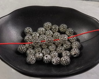 Può includere: Una collezione di perline sferiche in metallo color argento caratterizzate da un intricato motivo a reticolo traforato. Le perline sono raggruppate, con un singolo cordoncino rosso sottile infilato orizzontalmente al centro.