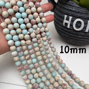 May include: Strands of 10mm round beads in shades of blue, beige, and red. The beads have a mottled, natural appearance. The image shows several strands of beads, held in a hand, with a plant pot in the background.