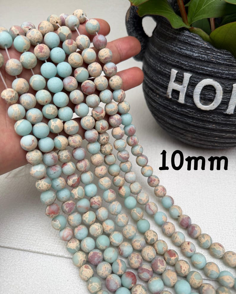 May include: Strands of 10mm round beads in shades of blue, beige, and red. The beads have a mottled, natural appearance. The image shows several strands of beads, held in a hand, with a plant pot in the background.