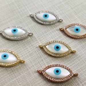 May include: Six eye-shaped charms with a white center, blue iris, and a surrounding border of small, clear stones. The metal frames are silver, gold, and rose gold. These charms are ideal for jewelry making.