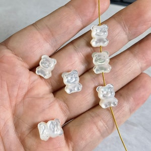 Puede incluir: Una colección de cuentas translúcidas con forma de osito de goma. Las cuentas son de un color pálido y nacarado y tienen un acabado liso y brillante. Están ensartadas en un fino alambre dorado, lo que sugiere que están destinadas a la fabricación de joyas o manualidades.