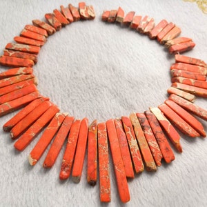 May include: A necklace composed of numerous rectangular, orange-red jasper beads. The beads have a natural, mottled pattern with tan and cream-colored veins. The necklace is arranged in a circular shape, showcasing the individual bead details.