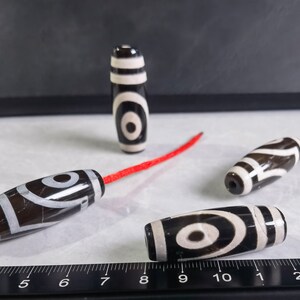 May include: Four black and white agate beads with a red string. The beads have a swirling pattern and a white eye design. The beads are on a white surface with a black ruler.
