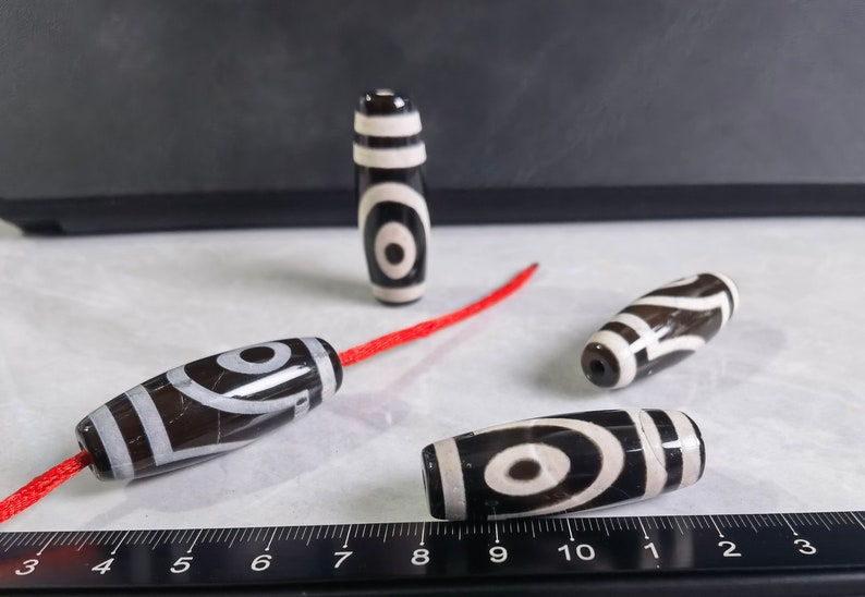 May include: Four black and white agate beads with a red string. The beads have a swirling pattern and a white eye design. The beads are on a white surface with a black ruler.