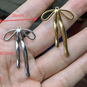 Op de afbeelding: Twee strikvormige metalen bedels, een zilverkleurige en een goudkleurige, elk met een klein lusje aan de bovenkant. Rode meetlijnen geven een breedte van 28 mm en een hoogte van 42,5 mm aan voor de zilveren bedel.